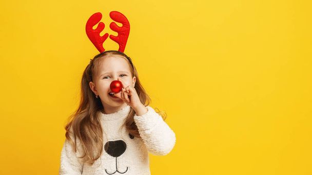 Happy little girl in fluffy white sweater with reindeer horns and a Christmas ball instead nose on yellow background. child is happy, having fun and preparing for Christmas. Advertising. Copy space. - Fotografie, Obrázek
