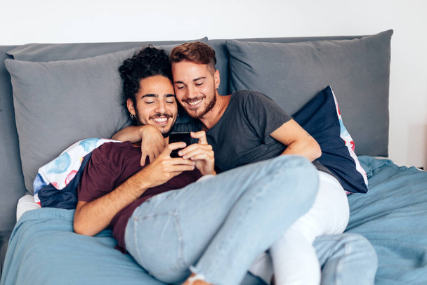 счастливая молодая гей-пара, лежащая на кровати и смотрящая видео на мобильном телефоне, или делая видеозвонок, или используя приложение - Фото, изображение