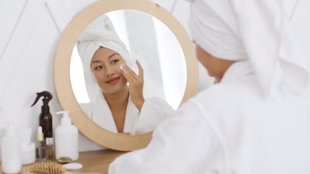 Bőrtisztító eljárások. Fiatal ázsiai hölgy tisztítja arcát testápolóval pamut pad, nézi a tükröt zuhanyzás után - Felvétel, videó