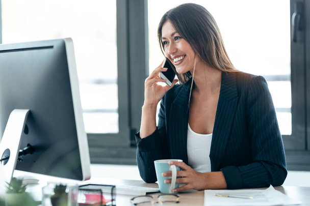 Shot van aantrekkelijke zakenvrouw praten met mobiele telefoon tijdens het werken met de computer in het kantoor. - Foto, afbeelding