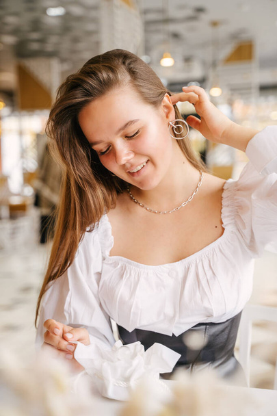 A beautiful girl sits in a cafe and nicely straightens her long hair behind her ear, showing off a beautiful silver earring - Valokuva, kuva