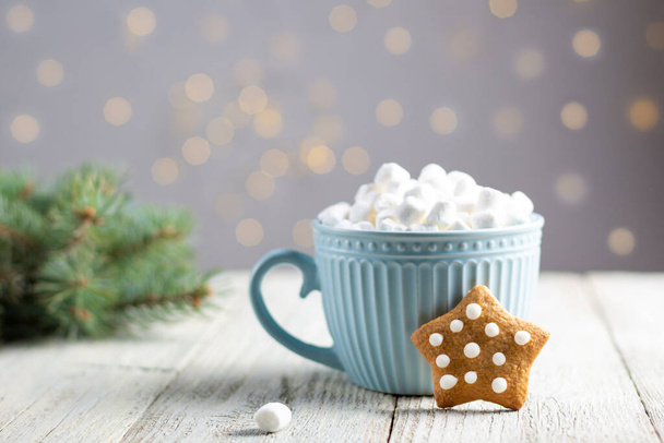 A blue cup of winter hot drink with marshmallows and gingerbread star on a white wooden background. Traditional Christmas atmosphere with bokeh. New year holiday concept. Festive background - Zdjęcie, obraz