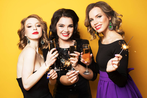 three young women with a champagne glasses and sparklers over yellow background - Valokuva, kuva