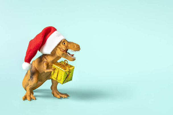 Dinosaur Rex in red Santa Claus hat holds golden gift box in its paws on blue background New Year Eve or Christmas Eve Art holiday card Creative idea for Merry Xmas concept. - Фото, изображение