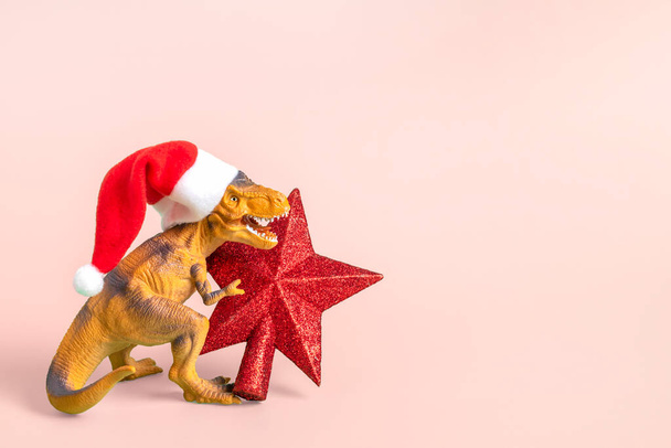 Dinosaur Rex in red Santa Claus hat holds star in its paws on pink background New Years Eve or Christmas Eve Art holiday card Creative idea for Merry xmas concept. - Photo, image