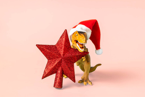 Dinosaur Rex in red Santa Claus hat holds star in its paws on pink background New Years Eve or Christmas Eve Art holiday card Creative idea for Merry xmas concept. - Photo, Image