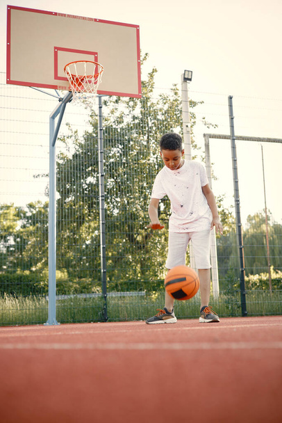 Мультирасовый мальчик стоит на баскетбольной площадке и играет с оранжевым мячом - Фото, изображение