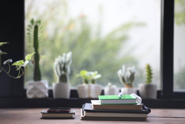 Блокноти з телефоном на дерев'яному столі та кактусі перед вікном
 - Фото, зображення