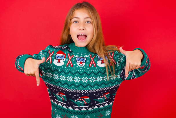 brunette kid girl in knitted sweater christmas over red background with positive expression, points down with both index fingers, has broad interested smile. Look there, please. - Photo, image