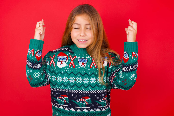 Χαρούμενη μελαχρινή κοπέλα με πλεκτό πουλόβερ Χριστούγεννα πάνω από κόκκινο φόντο σφίγγει τα δόντια, σηκώνει τα δάχτυλα σταυρώνει, κάνει επιθυμητή ευχή, περιμένει για καλά νέα, πρέπει να κερδίσω. - Φωτογραφία, εικόνα
