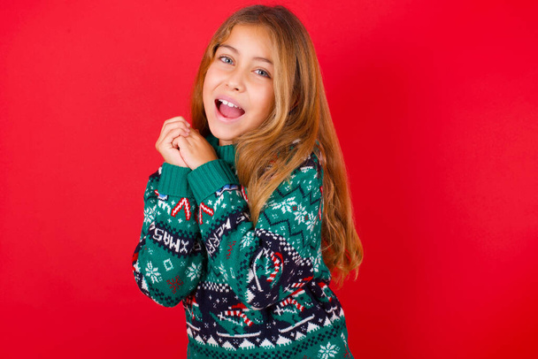 夢のようなブルネットの子供の女の子でニットセータークリスマス上の赤い背景で楽しい式、目を閉じて、手を顔の近くに交差させ続け、何か楽しいことについて考えて - 写真・画像