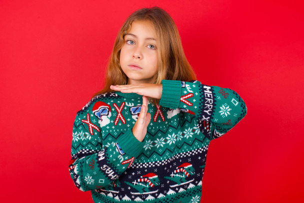 funny brunette kid girl in knitted sweater christmas over red background feels tired and bored, making a timeout gesture, needs to stop because of work stress, time concept. - Photo, Image