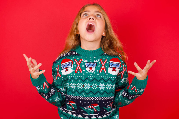 ニットセーターのブルネットの子供の女の子赤い背景に泣いて叫んでクリスマス。人間の感情、表情の概念。悲鳴、憎しみ、怒り. - 写真・画像