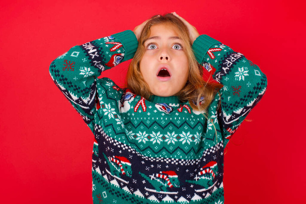 Horrible, stress, shock. Portrait emotional crazy brunette kid girl in knitted sweater christmas over red background clasping head in hands. Emotions, facial expression concept. - Фото, изображение