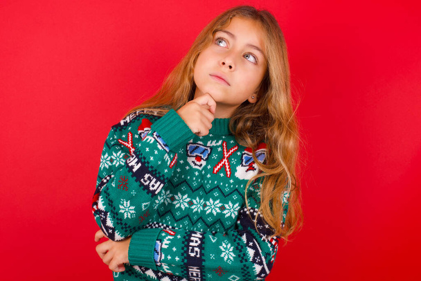 Výrazy tváře a emoce. Zamyšlená brunetka dítě dívka v pleteném svetru Vánoce přes červené pozadí drží ruku pod hlavou, s pochybovačným vzhledem. - Fotografie, Obrázek