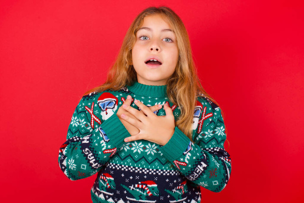 Gelukkig glimlachend brunette kind meisje in gebreide trui kerst over rode achtergrond heeft handen op de borst in de buurt van hart. Menselijke emoties, echte gevoelens en gezichtsuitdrukking. - Foto, afbeelding