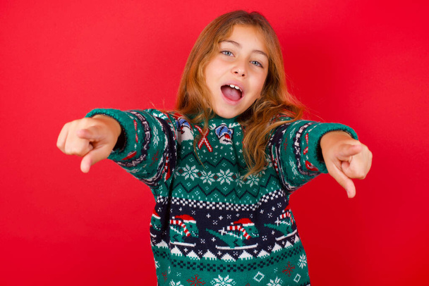 驚くべきブルネットの子供の女の子のクローズアップポートレートニットセータークリスマス赤い背景に2本の指でカメラを指していると言っ:私はあなたを選択! 、開く口で検索. - 写真・画像