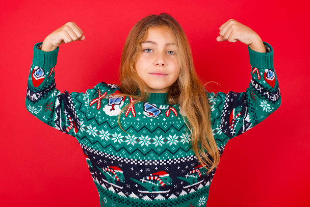 Vyötärö ylös laukaus brunette lapsi tyttö neulotut villapaita joulua yli punainen tausta nostaa kädet näyttää lihaksia tuntuu luottavainen voittoon, näyttää vahva ja itsenäinen, hymyilee positiivisesti kameralle. Urheilukäsite. - Valokuva, kuva