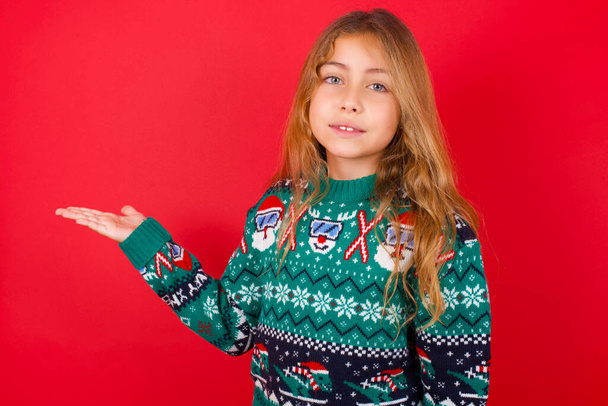 Positive glad brunette kid girl in knitted sweater christmas over red background says: wow how exciting it is, has amazed expression, shows something on blank space with open hand. Advertisement concept. - Zdjęcie, obraz