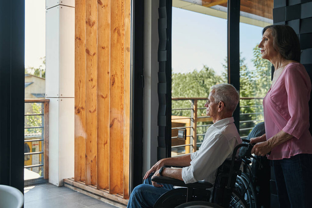 Femme âgée debout derrière l'utilisateur de fauteuil roulant masculin près de la fenêtre - Photo, image