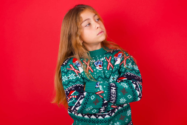 Image of upset brunette kid girl in knitted sweater christmas over red background with arms crossed. Looking with disappointed expression aside after listening to bad news. - Foto, Imagem