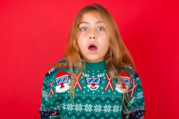 オープン口で赤い背景にニットセーターのクリスマスの感情的な魅力的なブルネットの子供の女の子は、カメラで大きな驚きと恐怖、星を表現しています。予期せぬ衝撃的なニュースと人間の反応. - 写真・画像