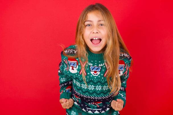 brunette kid meisje in gebreide trui kerst over rode achtergrond verhogen vuisten omhoog schreeuwen met vreugde zijn blij om doelen te bereiken. - Foto, afbeelding