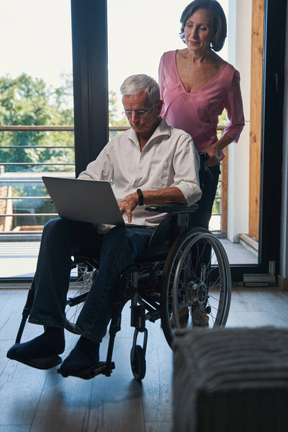 Aged woman pushing wheelchair with her husband working on laptop - Zdjęcie, obraz