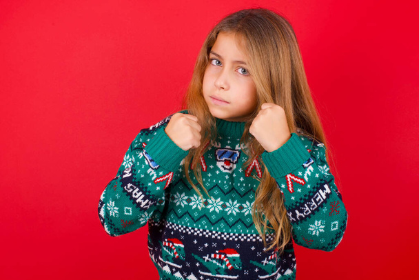 Portrét atraktivní brunetky dítě dívka v pleteném svetru Vánoce přes červené pozadí drží ruce před ním v boxerské pozici jít do boje. - Fotografie, Obrázek