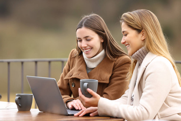 Two happy friends checking laptop in winter drinking coffee sitting in a terrace - Foto, imagen