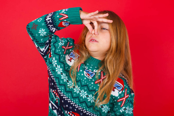 Ontevreden grappig brunette kid meisje in gebreide trui kerst over rode achtergrond pluggen neus als ruikt iets stinkt en onaangenaam, voelt afkeer, haat walgelijke geur. - Foto, afbeelding