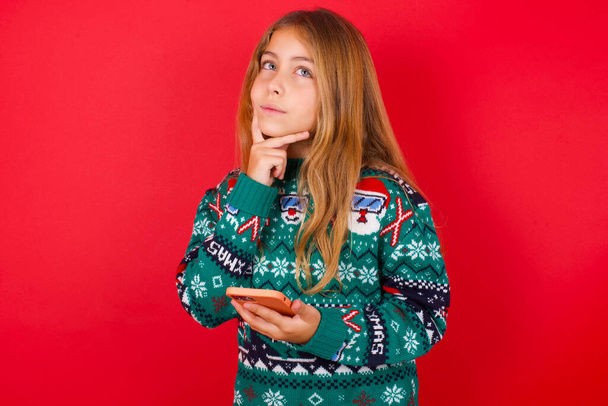 Σκέψη όνειρα αστεία μελαχρινή κορίτσι παιδί σε πλεκτό πουλόβερ Χριστούγεννα πάνω από το κόκκινο φόντο χρησιμοποιώντας το κινητό τηλέφωνο και κρατώντας το χέρι στο πρόσωπο. Λήψη αποφάσεων και έννοια των μέσων κοινωνικής δικτύωσης. - Φωτογραφία, εικόνα