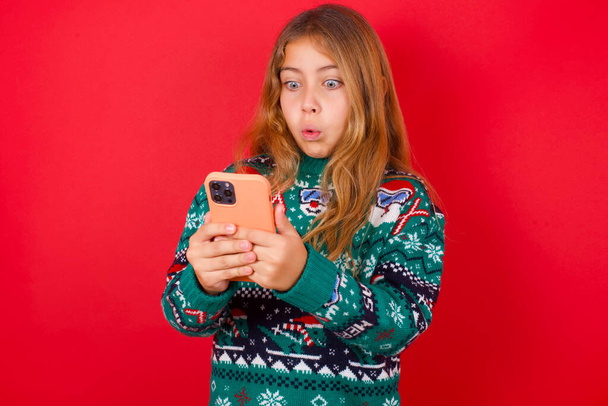 funny brunette kid girl in knitted sweater christmas over red background looks with bugged eyes, holds modern smart phone, receives unexpected message from friend, reads reminder. - Фото, зображення