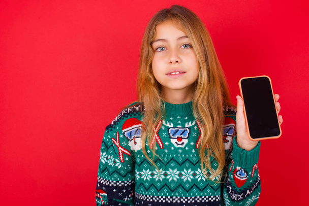 Χαμογελαστή μελαχρινή κοριτσάκι σε πλεκτό πουλόβερ Χριστούγεννα πάνω από το κόκκινο φόντο που δείχνει άδειο οθόνη του τηλεφώνου. Έννοια διαφήμισης και επικοινωνίας. - Φωτογραφία, εικόνα