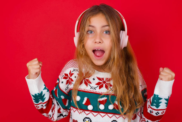 Emotional brunette kid girl in knitted sweater christmas over red background exclaims loudly feels like winner raises clenched fists keeps mouth opened wears stereo headphones on ears makes yes gesture, listens favourite music - Photo, Image
