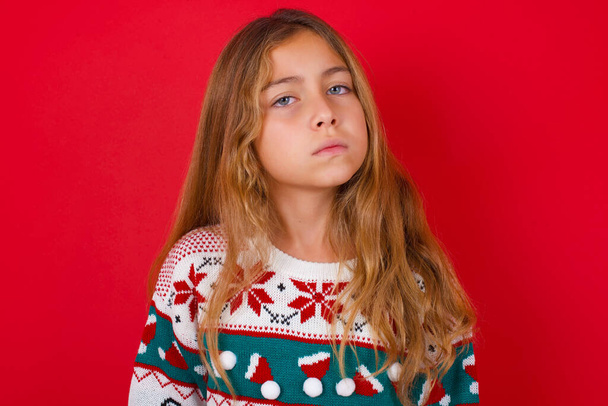 Menina criança morena descontente em camisola de malha Natal sobre vermelho fundo franjas rosto se sente infeliz tem alguns problemas. Conceito de emoções e sentimentos negativos - Foto, Imagem