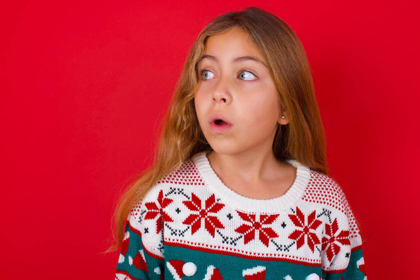 Zszokowana brunetka dziewczynka w swetrze na drutach Boże Narodzenie nad czerwonym tłem wyglądają puste miejsce z otwartymi ustami krzycząc: O mój Boże! Nie mogę w to uwierzyć. - Zdjęcie, obraz