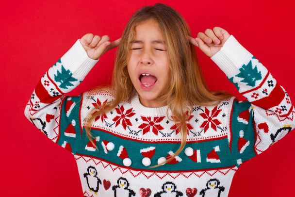 Fotografie šílené brunetky dítě dívka v pleteném svetru Vánoce nad červeným pozadím křičí a ukazuje prsty na vlasy zavřené oči - Fotografie, Obrázek
