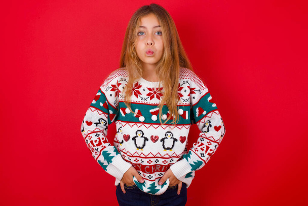 Portrait of lovely funny brunette kid girl in knitted sweater christmas over red background sending air kiss - Fotó, kép