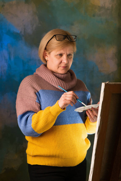 Artist paints a picture on canvas with oil paints. Works on his feet near the easel. Blonde in a blue and yellow sweater. Brushes and polish in hand. Focused and serious look. Painting masterpiece. Canvas is on an easel. Colored art background. - Photo, Image