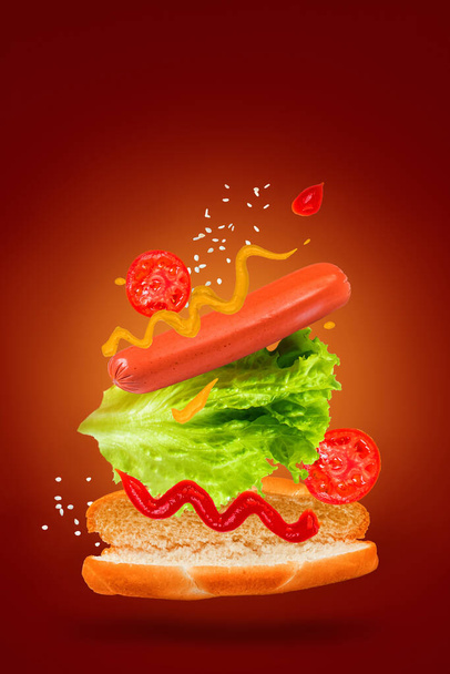 Fliegende Zutaten für einen leckeren Hot Dog auf rotem Hintergrund. Schwebewurst, Tomaten und Salat. - Foto, Bild