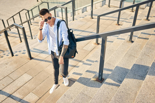 Ein junger Mann mit Sonnenbrille und schwarzem Rucksack steht auf der Treppe und lehnt am Geländer.. - Foto, Bild