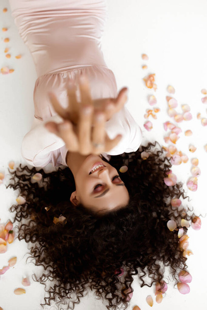 young caucasian woman in light dress in the white studio, a curly-haired european woman lies in rose petals , flowers in the hair - Фото, зображення