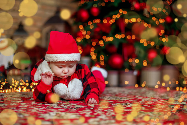 Een baby van zes maanden verkleed als kerstman. Achtergrond voor kerstkaart. kind kijkt neer op plaats voor inscriptie op de achtergrond van lichtgevende bloemenslingers en kerstboom. Kopieerruimte - Foto, afbeelding