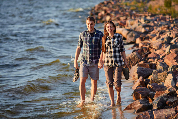 Feliz pareja caucásica de moda enamorada tomados de la mano y caminando por la costa cerca del río. En el fondo es la puesta de sol. - Foto, Imagen