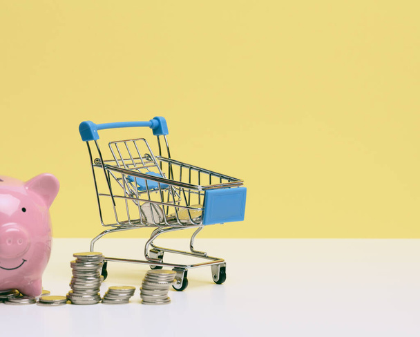 metal miniature shopping cart with change and pink piggy bank on a white table, yellow background. Concept of saving budget, discounts - Photo, Image