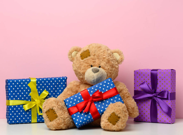 cute brown teddy bear holding a box wrapped in paper and red silk ribbon on white table. Prize and congratulations, pink background - Foto, imagen