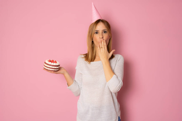 Joven mujer sorprendida con pastel de cumpleaños con signo de aprobación sobre un fondo rosa - Foto, imagen