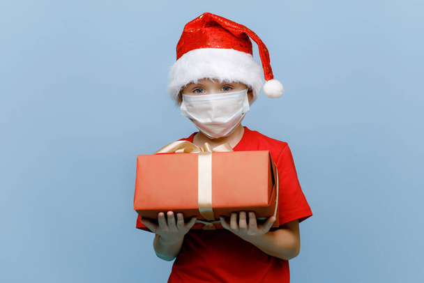 Portrait of a happy little boy in a medical mask, wearing a Christmas hat, holding a gift box and looking into the camera, isolated on a blue background. Copy space - Photo, image