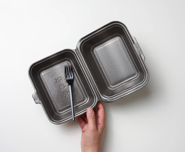 la mano femenina sostiene un recipiente de comida desechable y un tenedor de plástico. Caja gris de poliestireno en blanco sobre fondo blanco - Foto, imagen